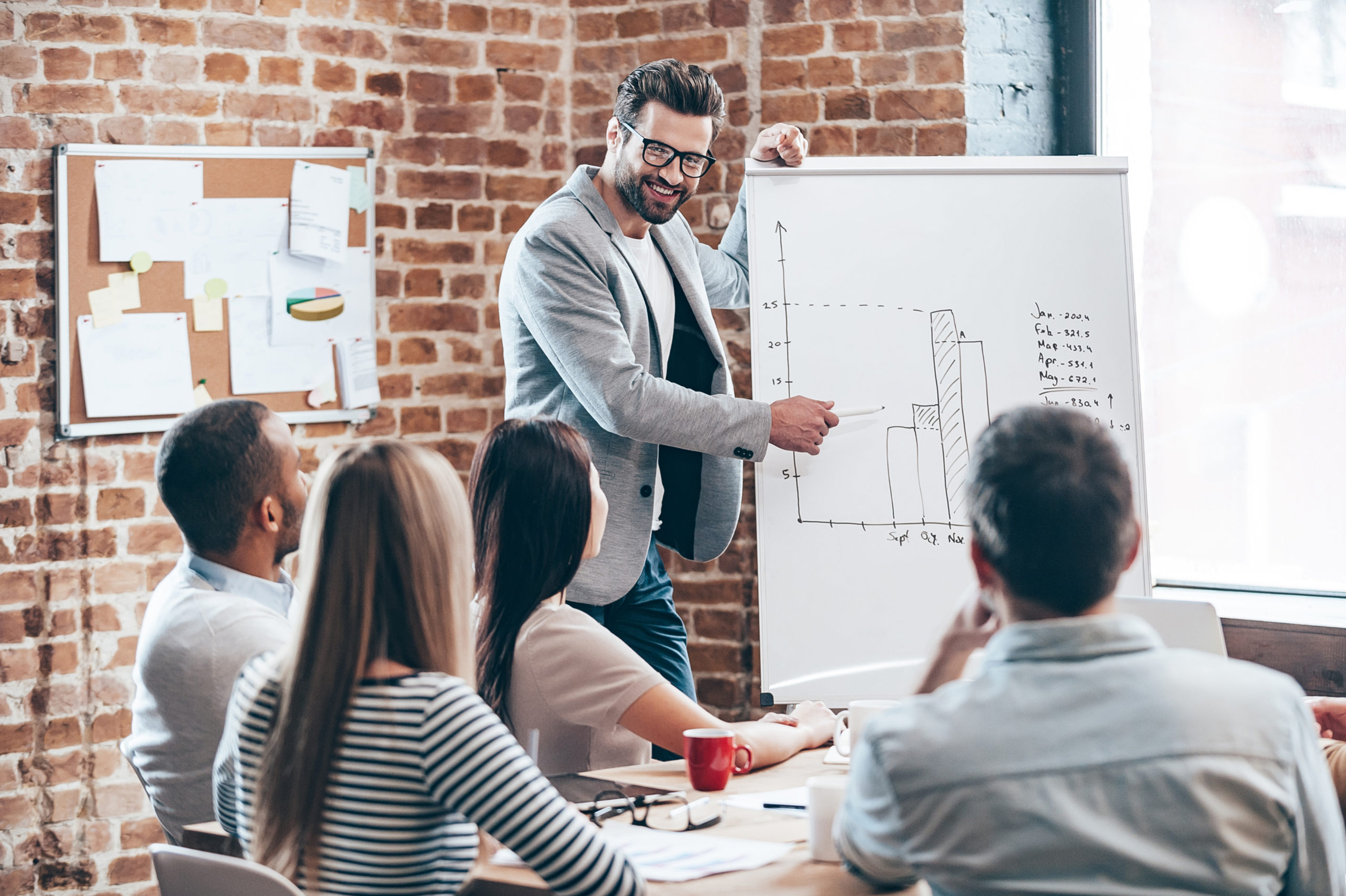 Look how fast growth our company! Handsome young man in glasses standing near whiteboard and pointing on the chart while his coworkers listening and sitting at the table